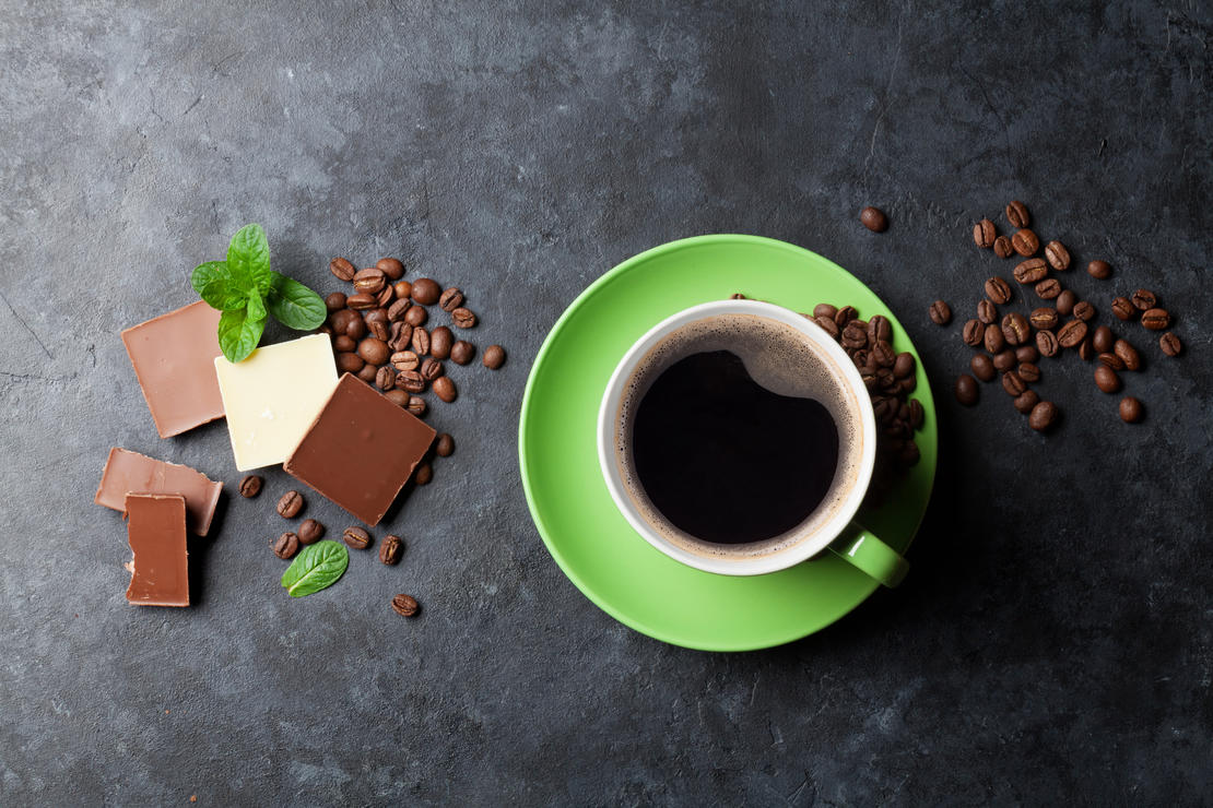 black coffee in a mug surrounded by pieces of dark, milk and white chocolate as well as coffee beans