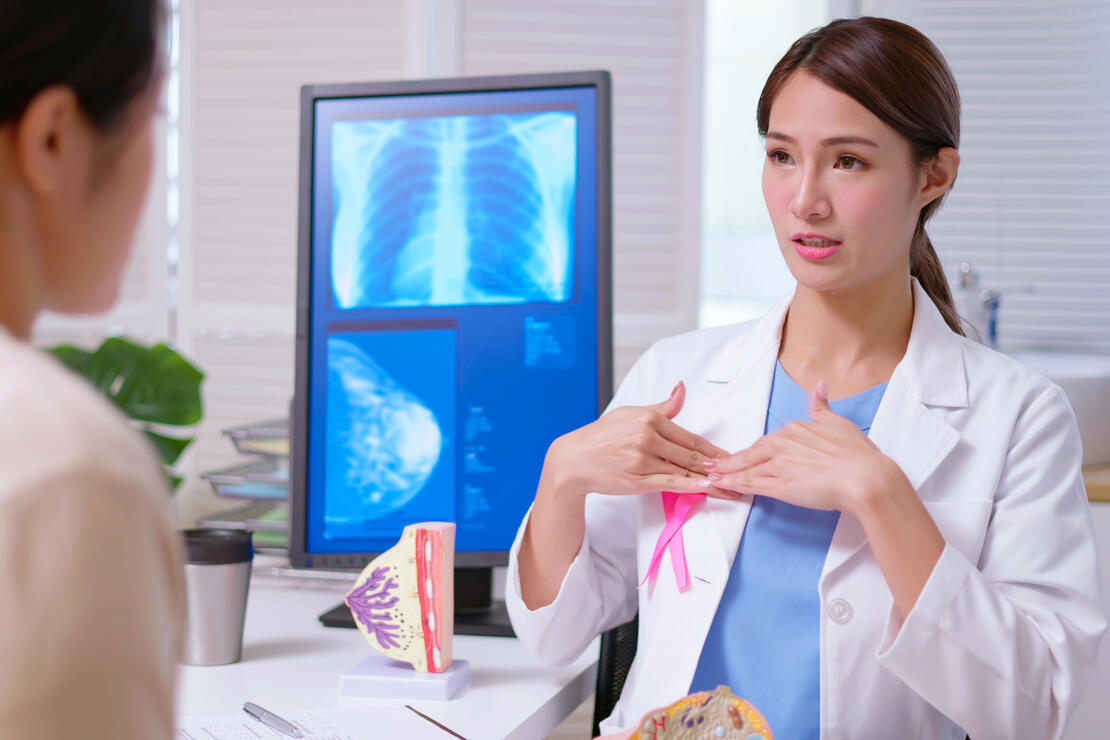 young female doctor wearing white coat with pink ribbon explaining breast self examination to woman at clinic - a model and xray display