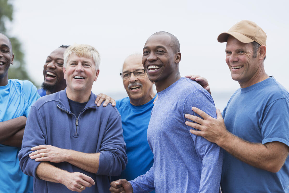 Multi-ethnic group of men standing together, smiling, supporting one another. Mixed ages ranging from 20s to 70s.