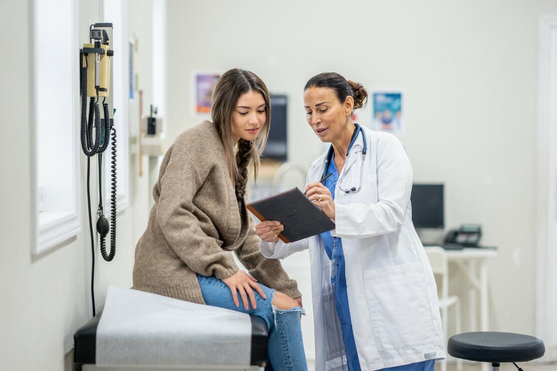 20-something female patient sitting on an exam table leans in to review her medical record or results, held out by her female physician