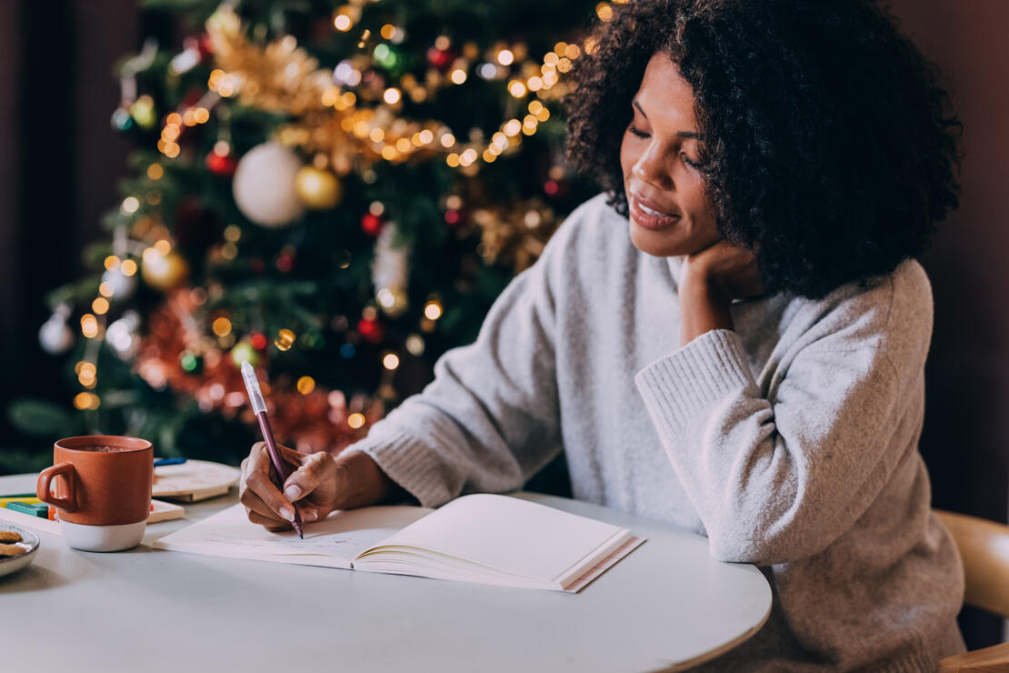 A smiling Latin female writing her New Year goals in her notebook while sitting at the table.