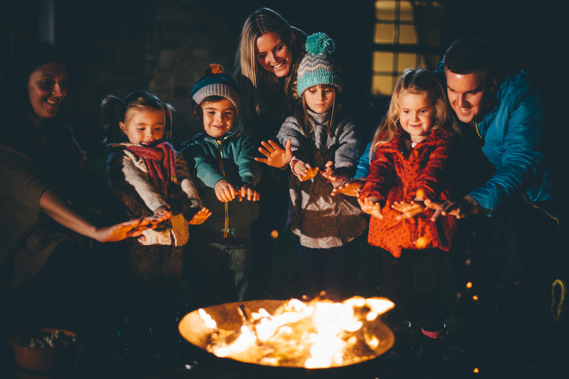 Group of family, friends and children gathered around a bonfire, holding their hands up to the fire to keep warm.