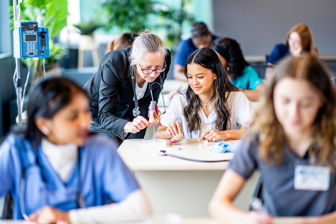 A large group of medical students are seen sitting in rows of desk as they work in pairs on various activities. They are each dressed professionally in scrubs and are working diligently on perfecting their skills. Their professor is seen checking in on a student in the background.