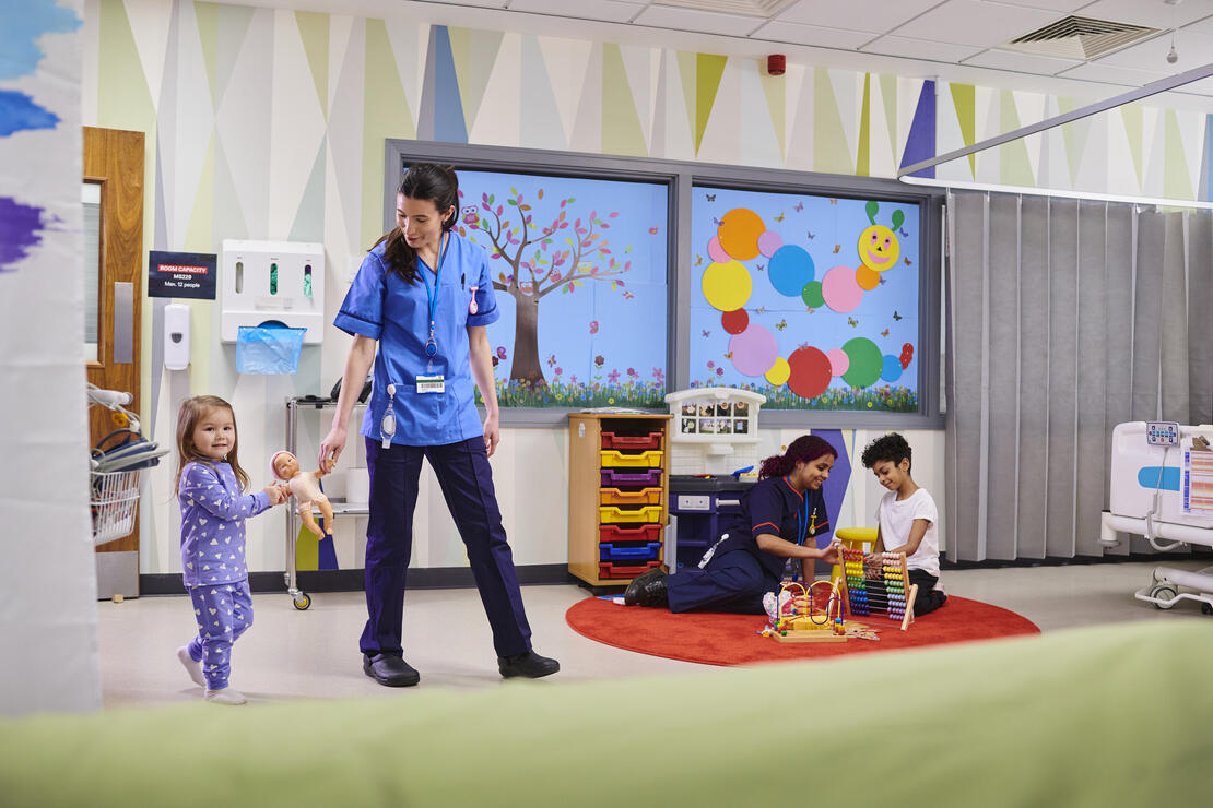 Little girl and her nurse walking, holding a baby doll between them. Another Child Life specialist plays with a young boy behind them.