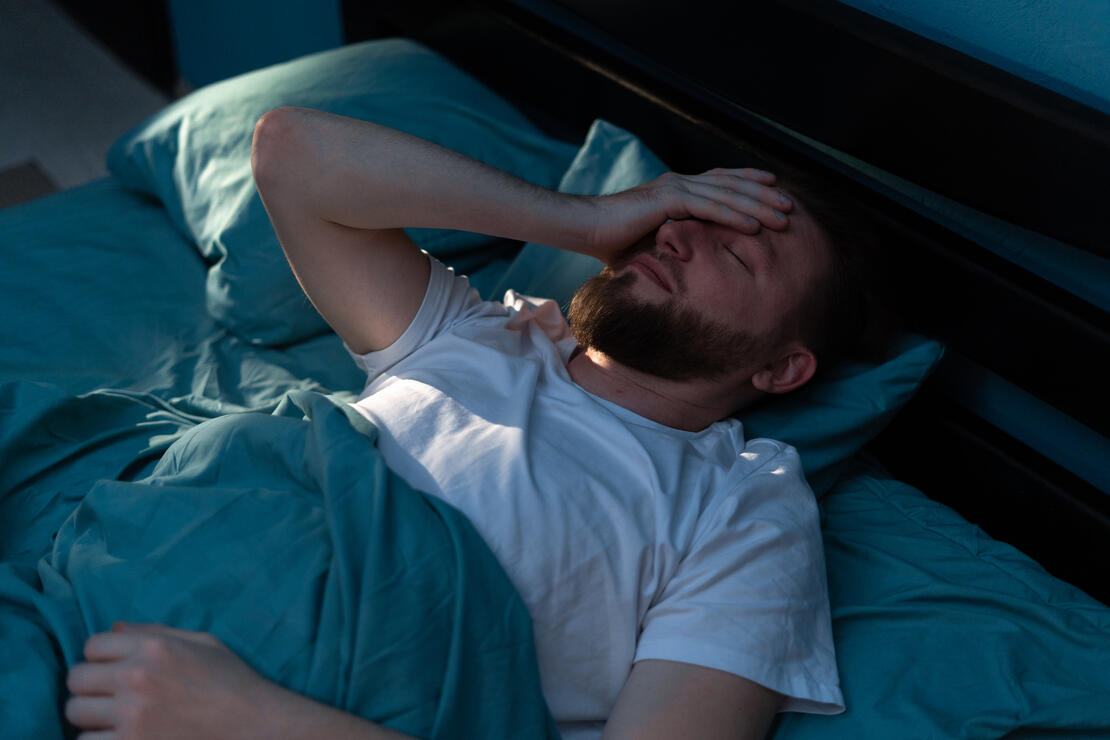 Man lying awake in bed, arm draped over his face with exhaustion.