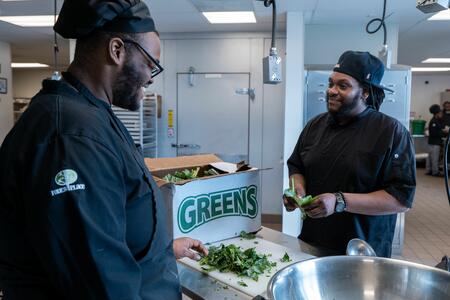Two chefs in black smiling over a cutting board and box of greens.