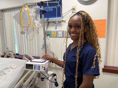 Photo of a nurse in blue scrubs smiling by an empty bed.
