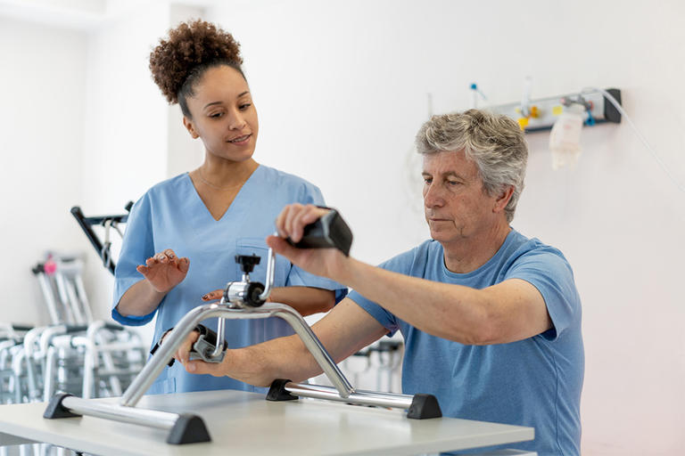 Black female therapist instructing senior patient how to exercise his arms on machine during physical therapy - Healthcare concepts