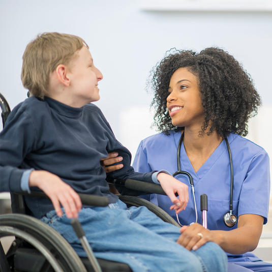 A nurse is helping a boy with cerebral palsy get into his wheelchair. They are both smiling and talking to each other.
