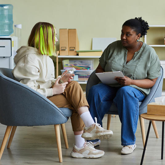 African female social worker talking to difficult teenage girl while they sitting on armchairs at office